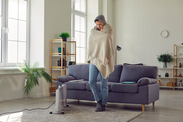 Obraz premium Man freezing inside residential building with no heating. Man feeling very cold at home, shivering, wearing plaid and winter hat, standing in living room interior with grey couch and electric heater