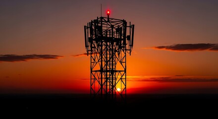 The silhouette of a cell phone tower stands against a dramatic sunset sky. The red warning light at the top of the tower is on.