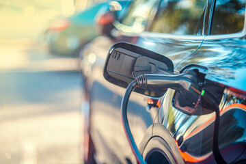 Electric car charging in sunny parking lot outdoors, focus on EV charger station cable plugged into charging port. Closeup.