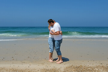 Couple in casual clothes embracing on a sandy beach near the ocean under a clear blue sky.