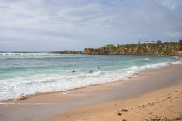 Group of surfers floating in sea near rocky shoreline. Peaceful beach with light waves and cloudy sky. Coastal scene capturing nature and outdoor water activity
