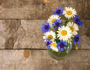 A charming bouquet of white daisies and vibrant blue cornflowers arranged in a clear glass vase on a rustic wooden table.