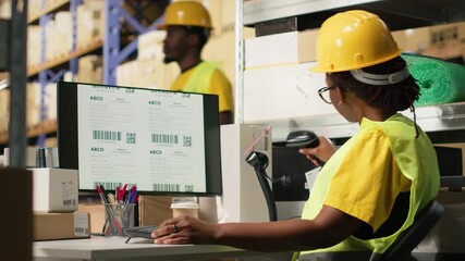 Black female with hi vis vest registering awb tracking info with scanner, oversees express delivery service for cargo orders. Scanning shipment details on labels, monitoring pc software. Camera B.