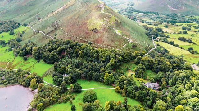 aerial panning right drone shot low angle of mountain with path leading up in Lake District England catbells with lake ullswater in view and nature and valleys surrounding in summer with cloudy skies