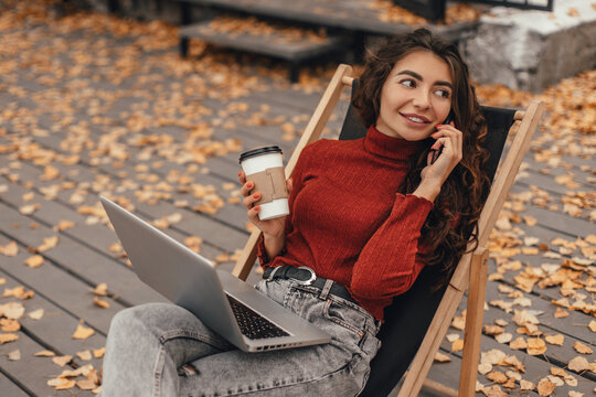 Woman in cozy outfit work at laptop and talk on the phone while sitting on chair near cafe in autumn park. Female student studies information using computer outdoor. Freelancer works remotely outside.