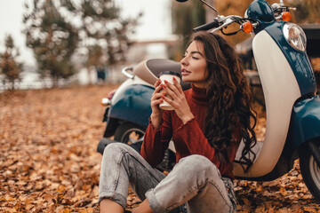 Beautiful woman in cozy sweater drink coffee or latte while sitting among the fallen leaves near vintage scooter in autumn park.