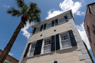 Charleston, South Carolina, USA - July 2, 2025: Low-angle view of a historic Charleston stucco house with black shutters and palm tree under a bright blue sky.