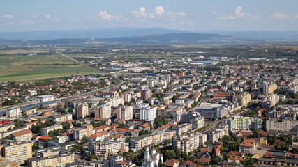 high-angle view of Deva, a city in Hunedoara County, Romania, showing a blend of old and new architecture, with lush green trees and a scenic backdrop