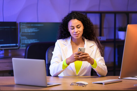 Young African-American QA engineer with mobile phone in office at night