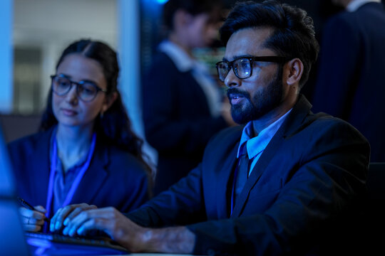 A diverse team of AI developers collaborates on a night coding session. A senior engineer oversees his team, intensely focused on debugging a complex machine learning project.
