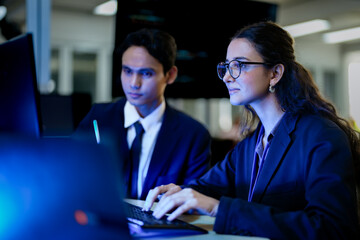 A diverse team of AI developers in a pair programming session. The female programmer and her male colleague collaborate, intensely focused on coding a complex new algorithm at night.