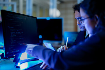 A diverse team of AI developers in a pair programming session. The female programmer and her male colleague collaborate, intensely focused on coding a complex new algorithm at night.