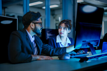 A diverse team of AI developers in a pair programming session. The female programmer and her male colleague collaborate, intensely focused on coding a complex new algorithm at night.