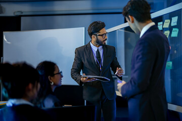 A senior AI architect leads a training session for a diverse team of developers. He explains a complex machine learning algorithm, pointing to code on a large vertical monitor.
