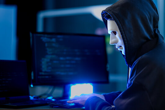 An anonymous hacker wearing a mask and hoodie in a dark room, launching a cyber attack. A concept for cybersecurity, data breach, ransomware, and internet crime.