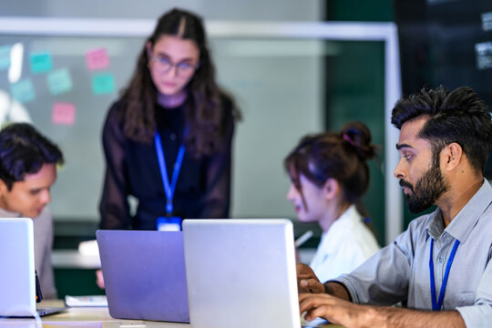 A diverse group of professionals in a corporate workshop on AI development. A female instructor supervises as the team learns to code advanced machine learning models on their laptops. - Powered by Adobe