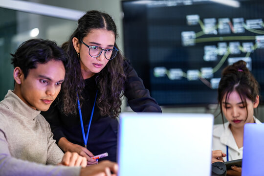 A diverse group of professionals in a corporate workshop on AI development. A female instructor supervises as the team learns to code advanced machine learning models on their laptops.