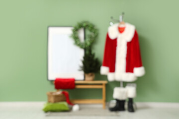 Fototapeta premium Hanger with Santa Claus suit, bench and frame near green wall. Blurred view