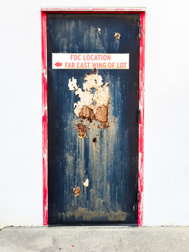 A worn and corroded red-framed blue metal door on a white building with a fire department connection sign