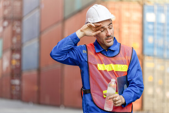 Tired Male Engineer in Safety Vest and Hard Hat Wiping Sweat at Shipping Container Yard, Overworked Freight Supervisor Taking a Break with Water Bottle Amidst Cargo Containers, Heat and Exhaustion