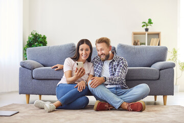 Happy positive husband and wife sharing phone screen at home together, sitting on warm floor in...