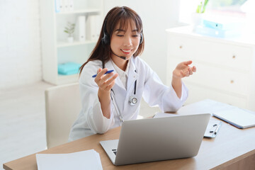 Female Asian doctor with headset and laptop giving online consultation in clinic