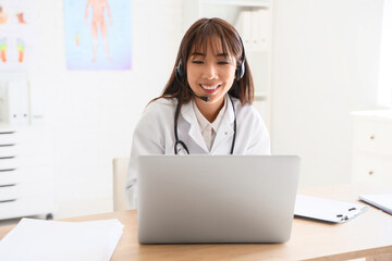 Female Asian doctor with headset and laptop giving online consultation in clinic