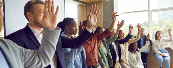 Portrait of a smiling joyful diverse company employees or a group of staff standing in a row in office raising their hands up to answer a question or voting on a business meeting. Banner.