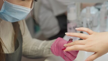 View of nail technician wearing pink glove and face mask carefully applying red nail polish to client fingernail with precision, surrounded by various manicure tools and cosmetics on white workspace