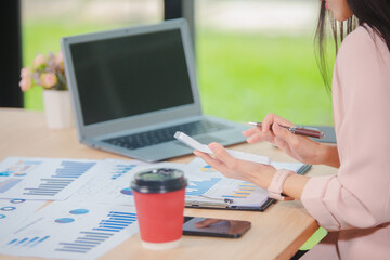 Smiling young Asian businesswoman is thinking and analyzing work plan in office hard using laptop, smartphone, calculator and documents. Chart sheet on table. Business and finance concept.
