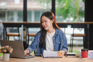 Smiling young Asian woman clearing paperwork in office, using laptop and many documents beside her. Accounting and finance business concept.
