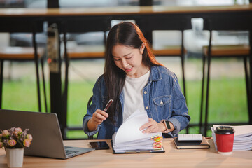 Smiling young Asian woman clearing paperwork in office, using laptop and many documents beside her. Accounting and finance business concept.