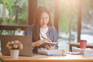 Smiling young Asian woman clearing paperwork in office, using laptop and many documents beside her. Accounting and finance business concept.