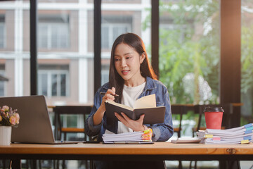 Smiling young Asian woman clearing paperwork in office, using laptop and many documents beside her. Accounting and finance business concept.