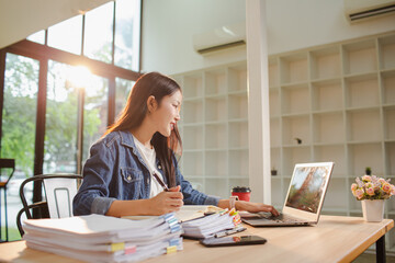 Smiling young Asian woman clearing paperwork in office, using laptop and many documents beside her. Accounting and finance business concept.