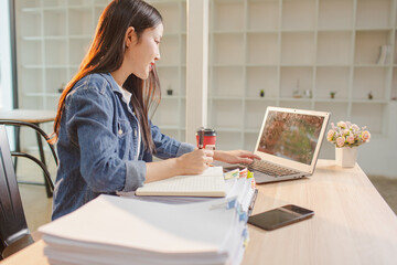 Smiling young Asian woman clearing paperwork in office, using laptop and many documents beside her. Accounting and finance business concept.