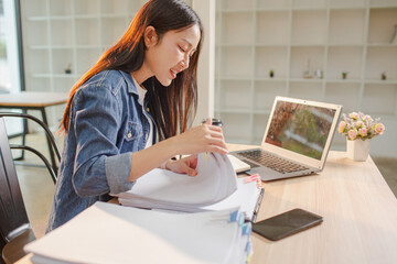 Smiling young Asian woman clearing paperwork in office, using laptop and many documents beside her. Accounting and finance business concept.