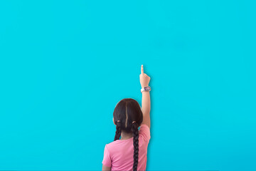 A girl points to a plain blue wall, empty space can placed for your text, mockup with children.