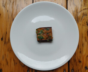 A top-down, minimalist view of a single piece of fried eggplant, topped with a garlic sauce and herbs. The piece is placed on a plain white plate, creating a lot of copy space, with the plate resting 