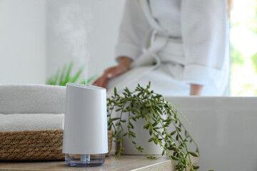 Air humidifier with towels and plant on table against woman sitting in bathroom, closeup