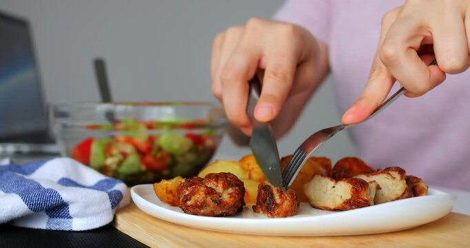 Hands using cutlery to cut and serve a portion of chicken meatballs and roasted potatoes from a white plate, with a bowl of fresh salad in the background