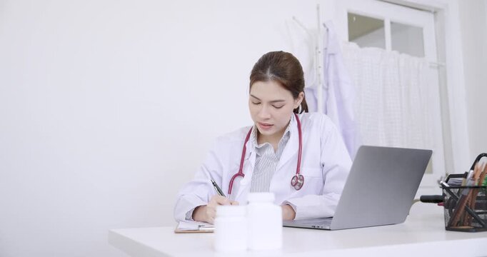 Caucasian female doctor sitting at desk writing medical notes during senior patient consultation in rehabilitation clinic with laptop and medicine bottles in clean clinical environment
