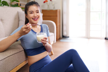 Asian young adult woman eating healthy yogurt bowl smoothie drink at home on floor beside sofa...