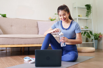 Asian adult woman engaging home fitness online workout session hydrating with water bottle using...