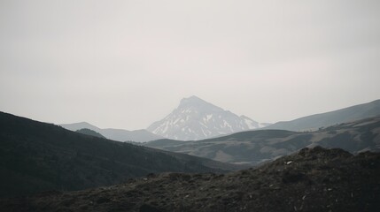 Majestic snow-covered mountain peak rising above dark rolling hills under a gray sky, capturing the serene beauty of winter wilderness landscape, perfect for nature background or travel inspiration.