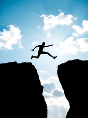 Silhouette of a man leaping across a gap between two cliffs against a bright blue sky with scattered clouds, symbolizing challenge, courage, and determination