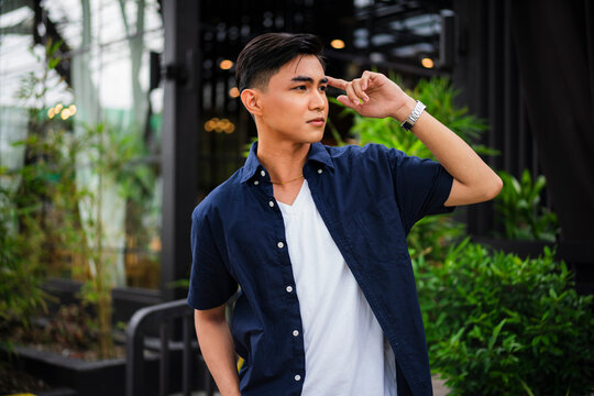 A confident young Asian man in a navy shirt and white tee poses with his hand raised to his temple outside a modern garden café.