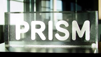 An illuminated clear block containing "PRISM" is captured in a well-lit indoor setting with a modern, reflective background - Powered by Adobe