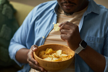 Young adult Black man sitting indoors holding wooden bowl eating potato chips with right hand...