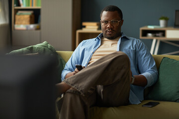 Portrait of Black man sitting on sofa holding remote control and watching television, wearing...